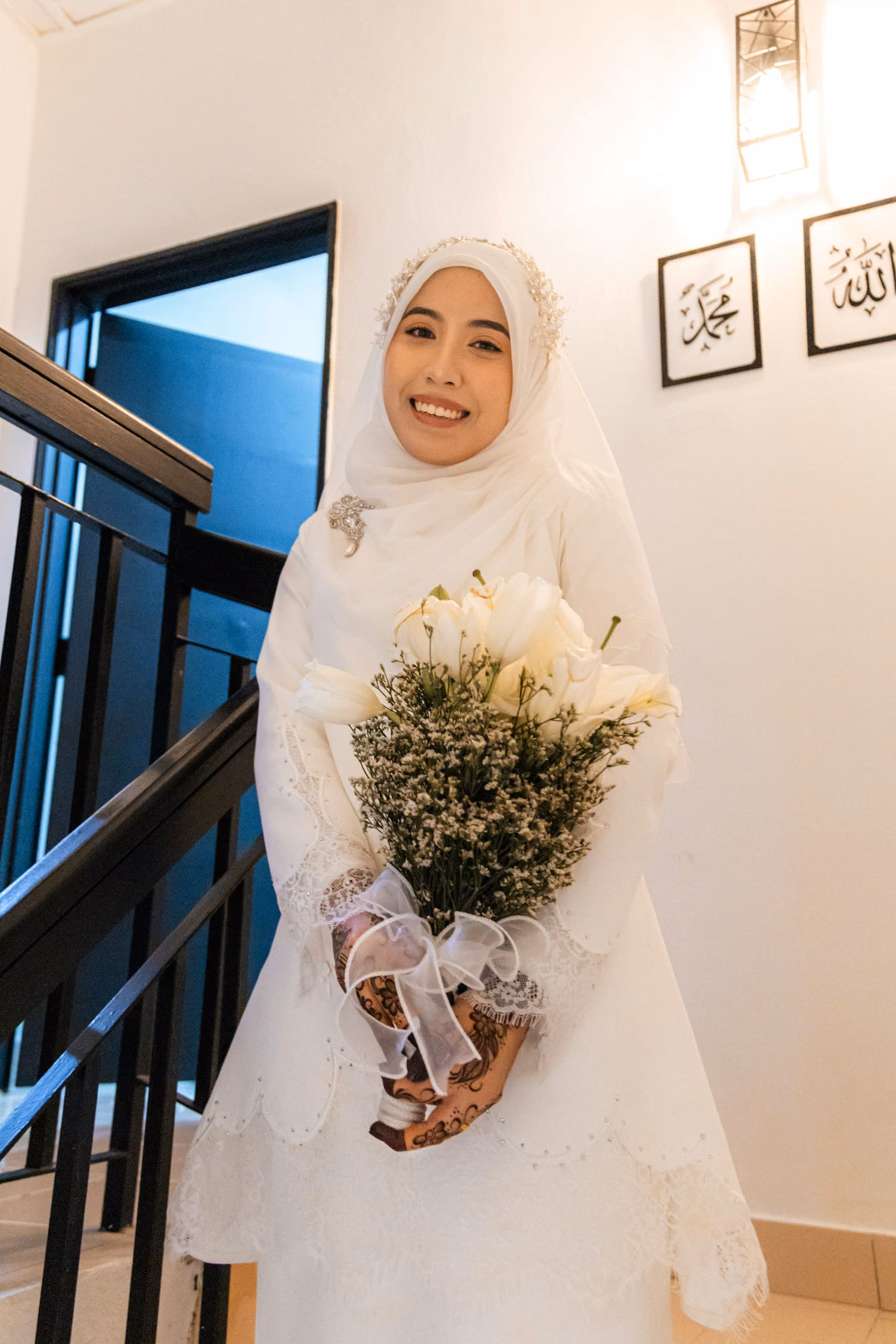 A bride in a white hijab and gown smiling on the stairs at home, holding a bouquet