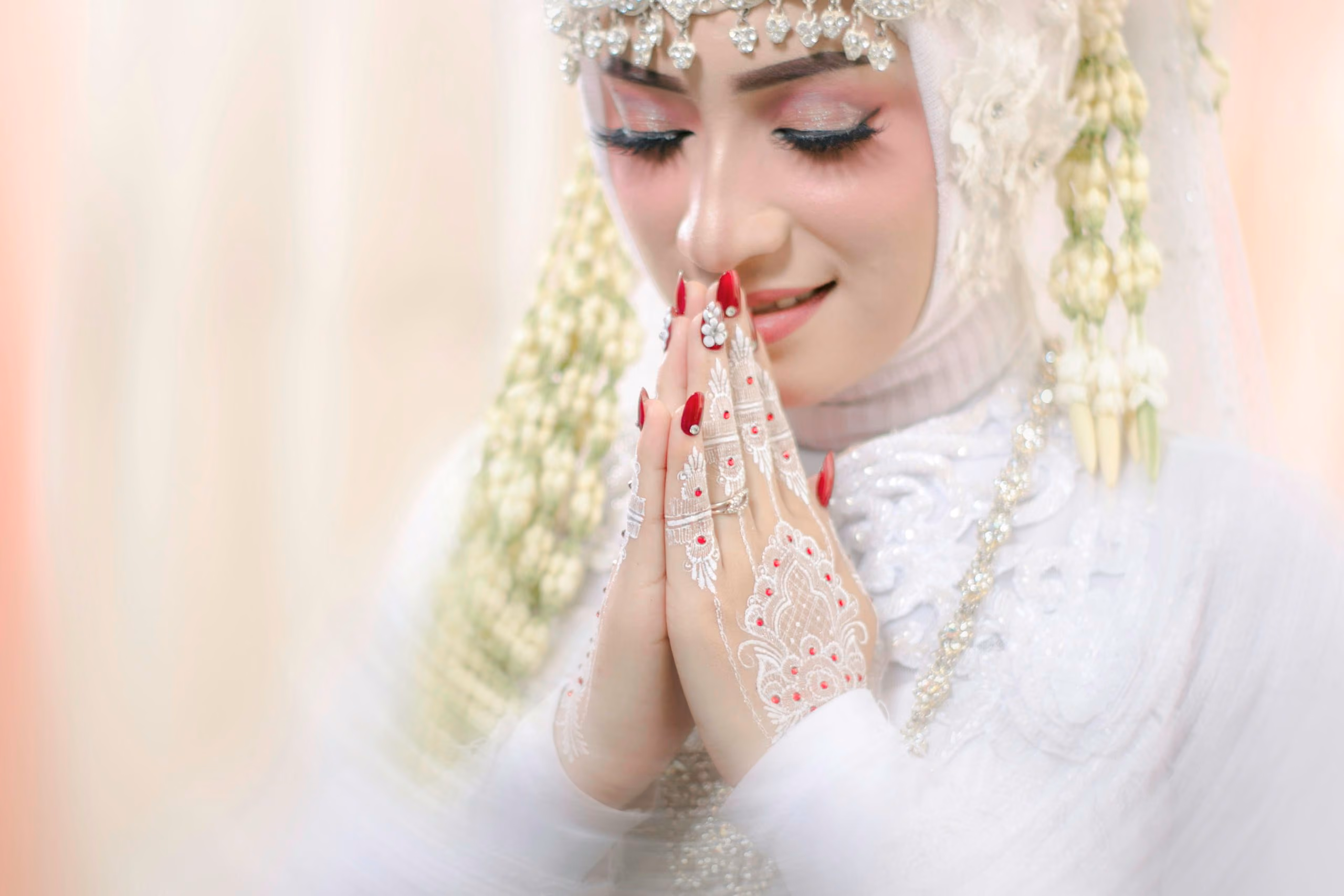 A bride in a white hijab with henna on her hands, eyes closed in prayer