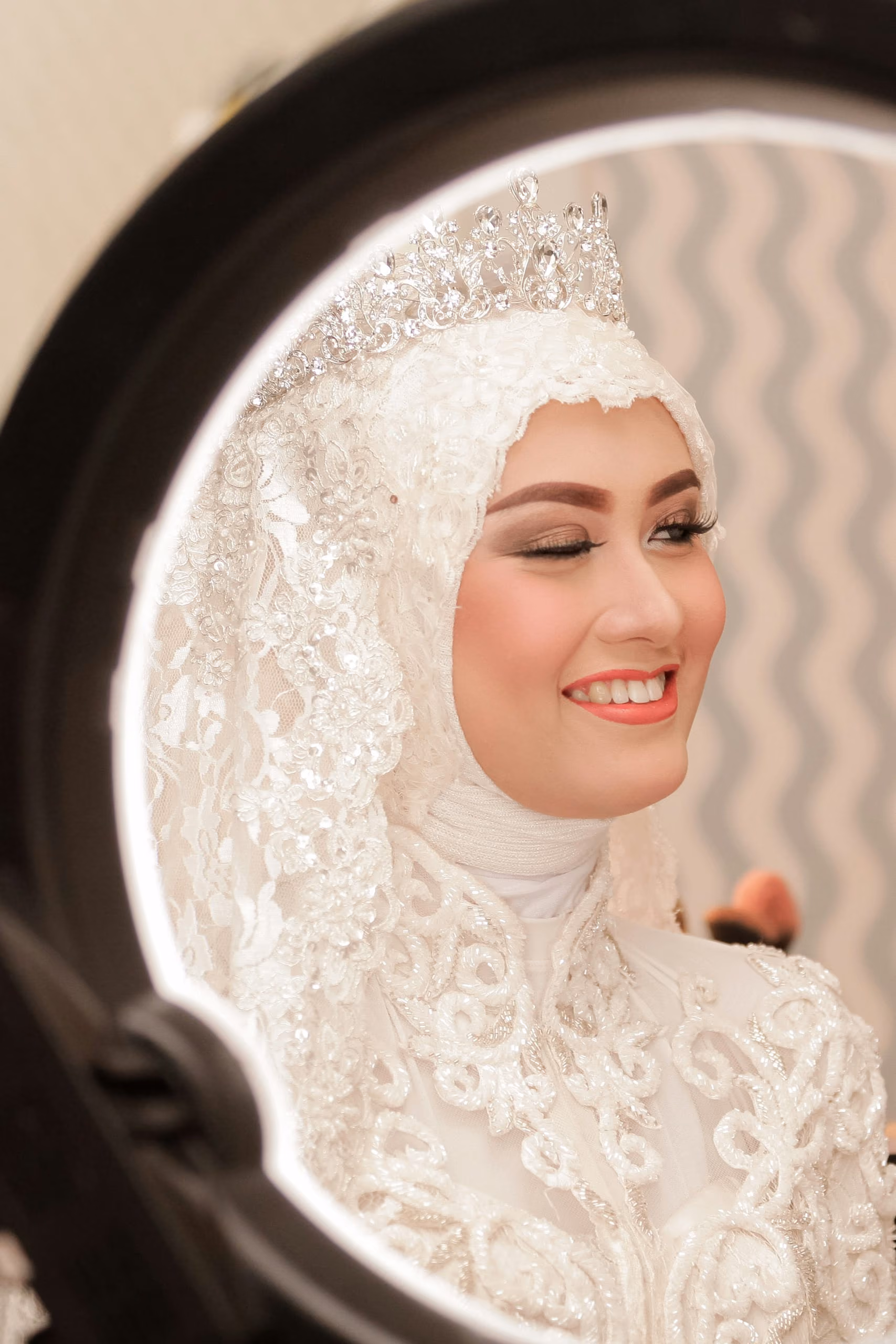 A bride in a white hijab and tiara smiling at her reflection in a round mirror