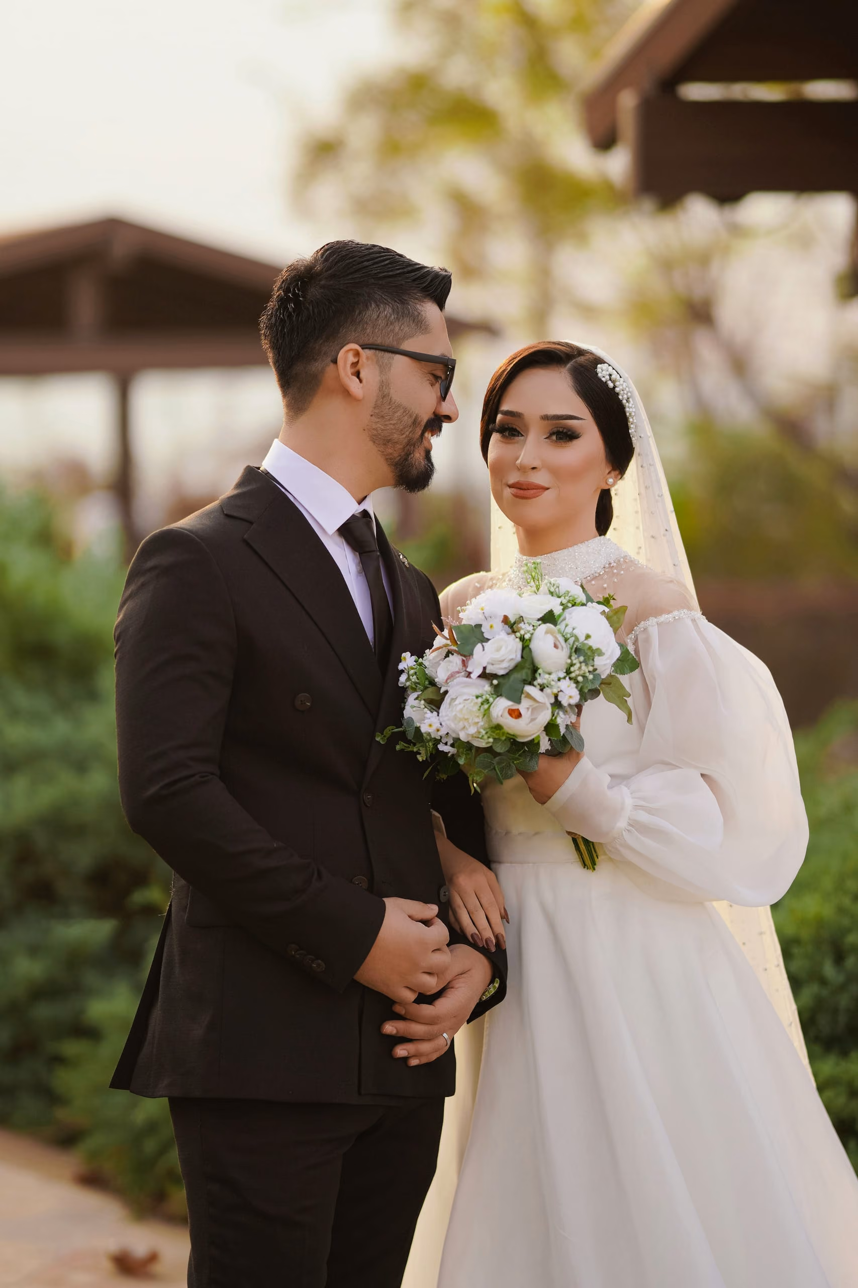 A bride and groom smiling at each other in a garden, she holds white roses