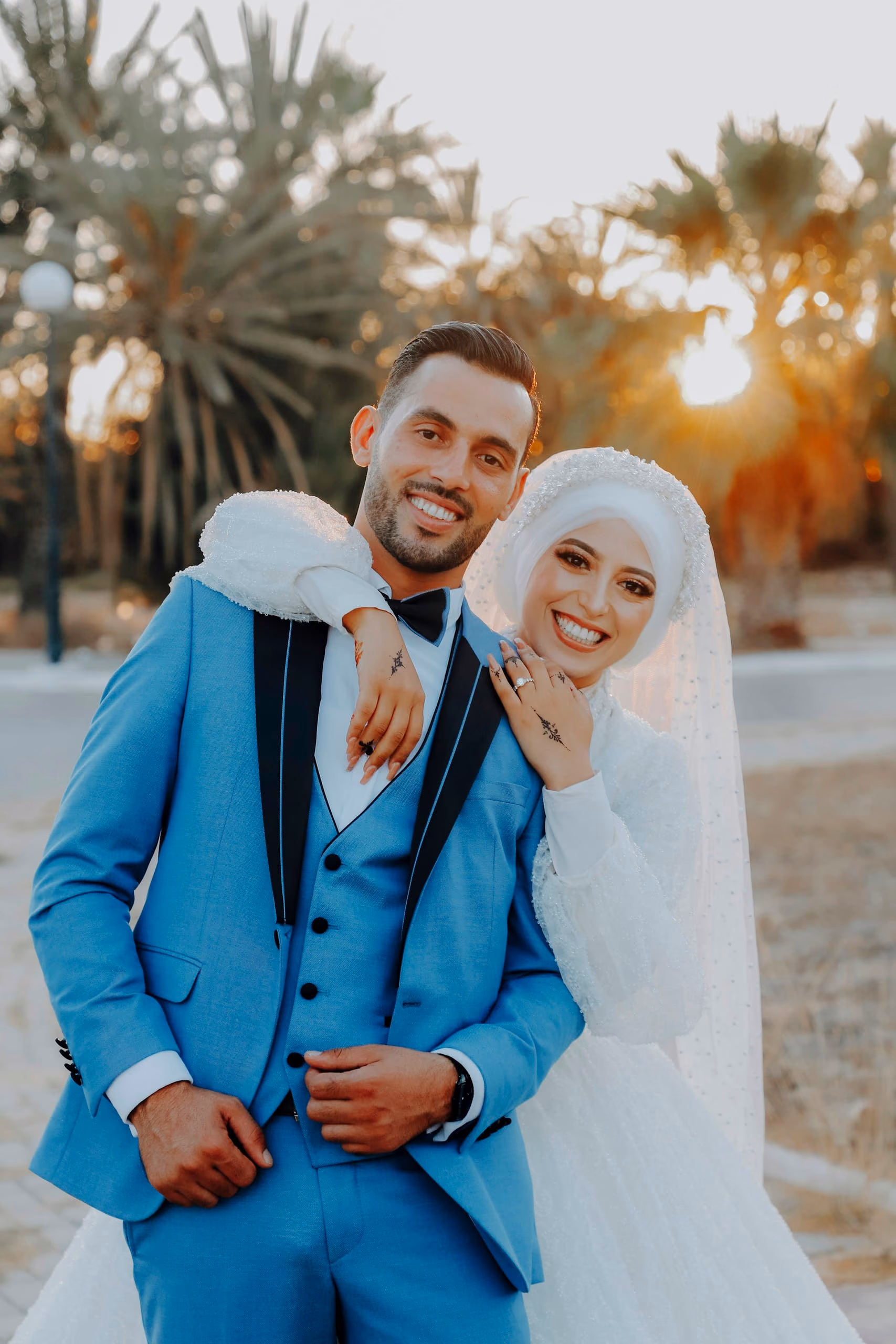 A groom in a blue suit and a bride in a white hijab laughing at sunset